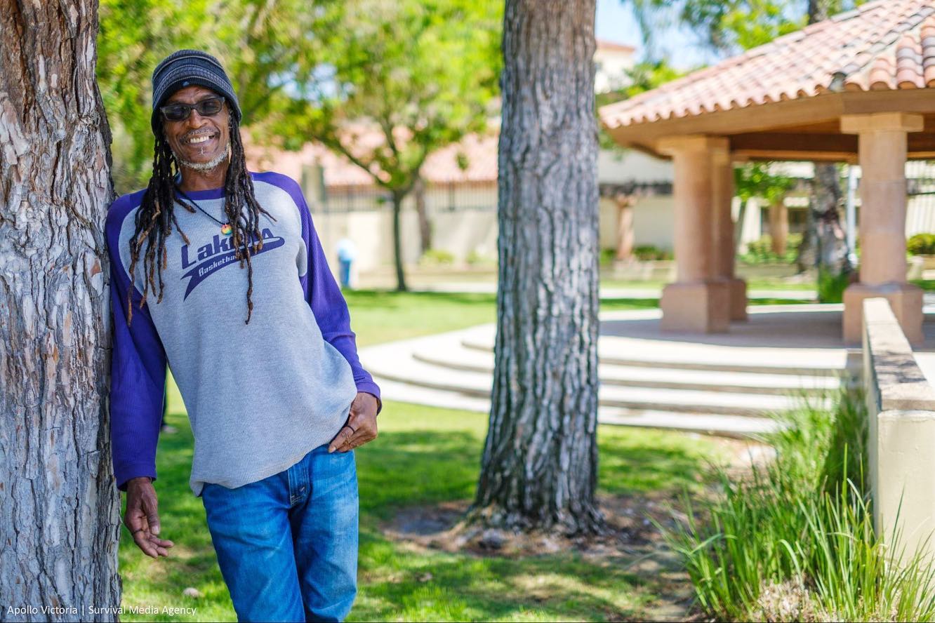 Picture of Brian Bukle, a middle aged Black man with long hair leaning against a tree, wearing a beanie. The image is brightly lit and in the background is green grass, leaves, and another tree.